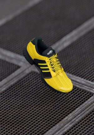 Żółty but sportowy z teksturowaną powierzchnią, czarnymi akcentami i trzema czarnymi paskami. Posiada wyściółkę amortyzującą i elastyczną podeszwę.