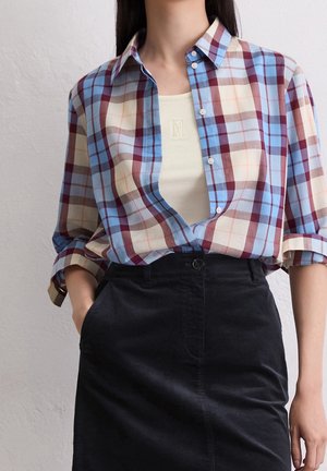 Vrouw draagt een blauw, rood en beige geruit overhemd over een witte tanktop, gecombineerd met een zwarte high-waist rok, met één hand in de zak.
