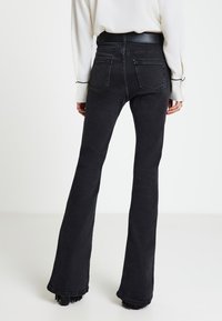 Persona che indossa jeans neri a vita alta con gamba svasata e tasche posteriori, una camicetta bianca a maniche lunghe e scarpe nere con tacco.