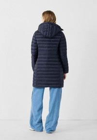 Marineblauwe gewatteerde jas met een capuchon, voorzien van een horizontaal gequilt design, lange mouwen en een gemiddelde lengte. Draagt over lichtblauwe wijde jeans.