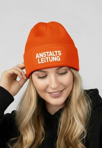 Orange knit beanie featuring a white rectangular patch with the text "ANSTALTS LEITUNG." Ribbed texture, snug fit, and rounded top design.