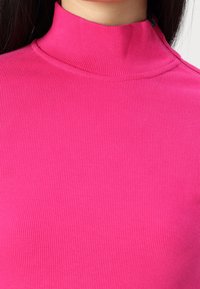 Přiléhavý rolák v živé růžové barvě, s ribkovanou strukturou, vysokým límcem a jemnými švy, vyrobený z měkké elastické tkaniny.