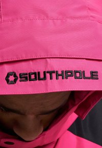 Rosa vanntett jakke med kontrasterende svart søm på kragen. Ordet "SOUTHPOLE" er brodert tydelig i svart. Jevn, matt tekstur.