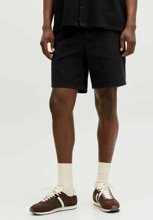 Hombre con pantalones cortos negros, calcetines blancos hasta el tobillo y zapatillas marrones con rayas blancas, de pie sobre un fondo claro.