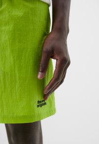 Lime green shorts with textured fabric; features a black logo "the new originals" near the hem; hand positioned above the shorts.