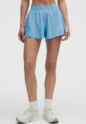 Femme portant un short de sport bleu clair, des chaussettes blanches et des chaussures de course blanches, debout devant un fond clair uni.