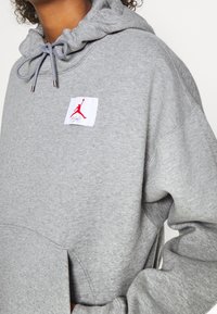 Felpa grigia con cappuccio, dotata di tasca frontale e di un patch del logo Jordan rosso. Tessuto strutturato con dettagli in cordoncino al collo.
