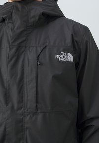 Veste imperméable noire avec capuche, poche zippée sur la poitrine et logo blanc "The North Face" sur le côté gauche de la poitrine.