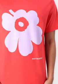 Červené bavlněné tričko s velkou grafikou levandulového květu a bílým logem "marimekko" blízko spodního okraje, umístěné na jednobarevném pozadí.