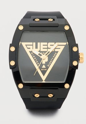 Montre-bracelet Guess noire et or avec un cadran carré, des vis dorées sur la lunette et un logo triangulaire audacieux sur le cadran.