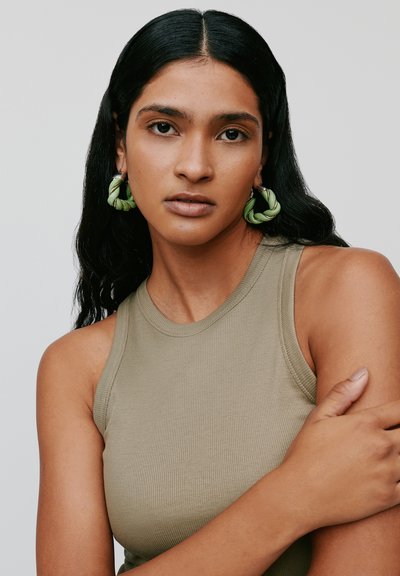 Top senza maniche in tessuto a coste color khaki, abbinato a orecchini a cerchio verdi intrecciati. La modella posa con le braccia incrociate, capelli ondulati di lunghezza media.