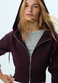 Felpa corta color bordeaux con zip completo e cappuccio con coulisse, dotata di un orlo a coste e dettaglio in metallo argentato sulla cerniera, indossata sopra una maglia grigia.