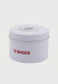 Rund hvid metalbeholder med rød "G-SHOCK" tekst og et cirkulært 30-års jubilæumslogo på låget.