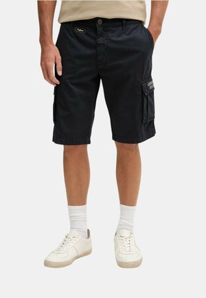 Uomo che indossa pantaloncini cargo neri con tasche laterali, camicia beige infilata, calzini bianchi e sneakers bianche, in piedi con le mani in tasca.