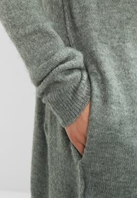 Hand delvis inskjuten i fickan på en lång, mjuk, grå, stickad tröja med ribbad mudd på ärmen.