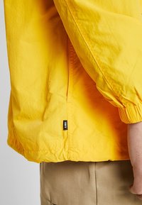 Yellow windbreaker jacket with elastic cuffs, zip side pocket, and a small black label. The fabric appears lightweight and slightly textured.