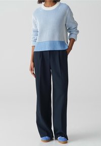 Person som bär en blå och vit randig tröja, vida svarta byxor och blå sneakers, står med ena handen i fickan.