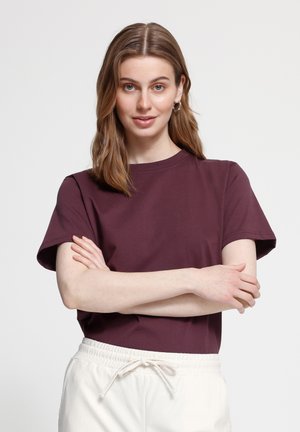 Jeune femme aux cheveux châtain clair, portant un t-shirt manches courtes bordeaux et un pantalon blanc à cordon, debout les bras croisés.