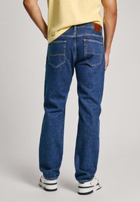 Jeans de mezclilla azul con un diseño de pierna recta, cinco bolsillos, costuras visibles y un parche de cuero marrón en la parte posterior de la cinturilla.