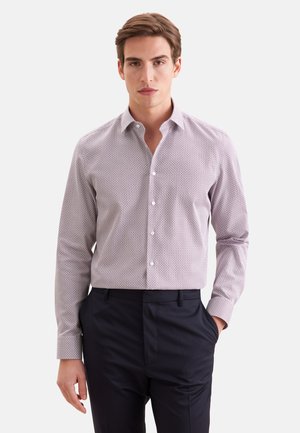 Jeune homme portant une chemise claire à manches longues à motifs, rentrée dans un pantalon noir, avec une main dans la poche, debout devant un fond uni.
