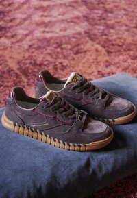 Par de zapatillas de ante marrón con suelas de goma y detalle de costura negra, colocadas sobre un cojín azul sobre una alfombra rojiza.