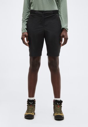 Άτομο που φοράει μαύρο σορτς μέχρι το γόνατο, πράσινο μακρυμάνικο μπλουζάκι, λευκές κάλτσες και χακί ορειβατικά μποτάκια, στέκεται σε λευκό φόντο.