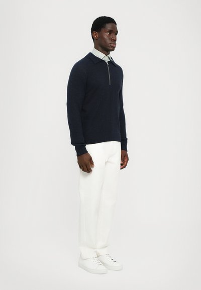 Suéter de punto azul marino con cremallera de cuarto y cuello con solapa, combinado con pantalones blancos ajustados y zapatillas blancas. Diseño simple y moderno.