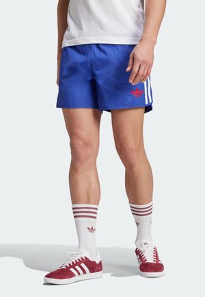 Mężczyzna noszący niebieskie szorty Adidas z białymi paskami i czerwonym logo, białe skarpety z czerwonymi paskami oraz czerwone buty sportowe Adidas z białymi sznurowadłami.