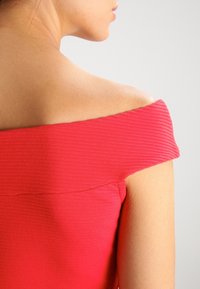 Czerwona bluzka typu off-the-shoulder o prążkowanej strukturze, z szerokim dekoltem i gładkim materiałem. Skóra widoczna przy dekolcie i ramieniu.