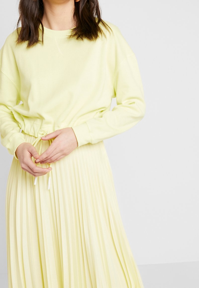 Felpa gialla chiaro con una vestibilità rilassata e maniche lunghe, abbinata a una gonna plissettata coordinata con vita regolabile.
