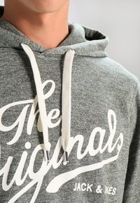 Felpa grigia mélange con logo stampato bianco, cordini con estremità metalliche e una texture morbida. Presenta un cappuccio e una vestibilità comoda.