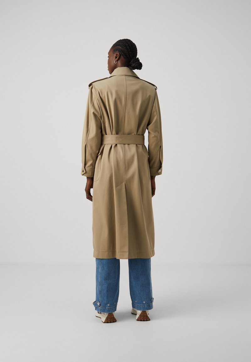 Langer beiger Trenchcoat mit Gürtel in der Taille, Schulterklappen und geknöpften Manschetten, kombiniert mit hellblauen weit geschnittenen Jeans.