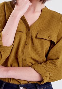 Personne à la peau claire portant une chemise boutonnée texturée moutarde avec des manches retroussées, la main près du visage, et un jean en denim bleu foncé.