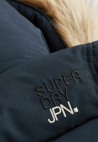 Tamno plavi puffer kaput s krznom na kapuljači. Ima izvezeni logo "SUPER DRY JPN" u crnoj i bijeloj boji na rukavu. Glatka tekstura.