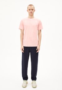 Lichtroze katoenen T-shirt met korte mouwen, gecombineerd met donkerblauwe broek. Model draagt meerkleurige sportschoenen. Minimalistisch ontwerp.