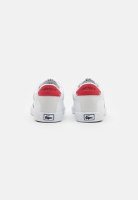 Baskets en cuir blanc avec un accent rouge sur le talon portant le logo "LACOSTE" et des surpiqûres détaillées, complétées par un logo bleu marine sur la semelle.
