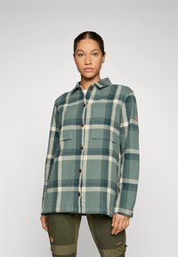 Camicia in flanella a quadri verde e crema con colletto, bottoni frontali, due tasche sul petto e una sottile etichetta con logo sulla manica.