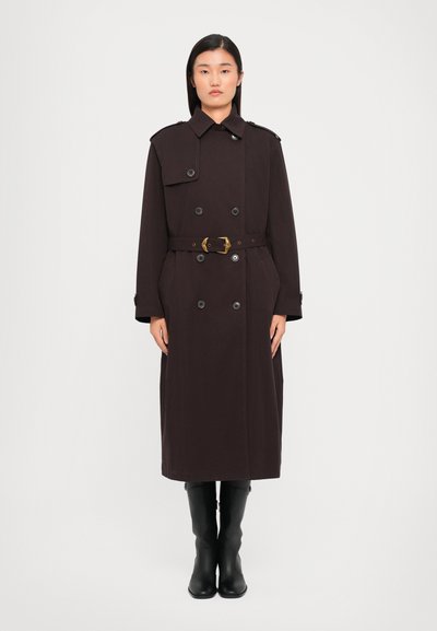 Trench coat doppiopetto in tessuto marrone scuro, con vita accentuata da una cintura, spalle con pattine e tasche laterali. Stivali neri al ginocchio.