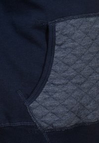Felpa blu navy con tasca grigia trapuntata. Presenta polsini e fondo a coste. Tessuto morbido con una finitura liscia.