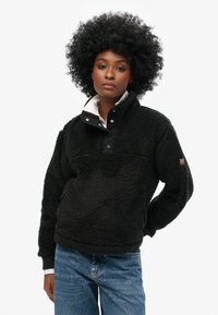 Pull en polaire noir avec un col montant, fermeture à boutons-pression et tissu texturé. Présente un col intérieur blanc et un logo brodé sur la manche.