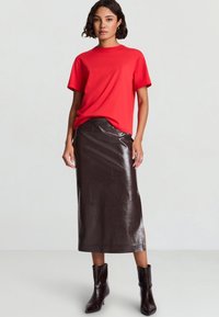 Maglietta rossa a maniche corte, abbinata a una gonna in pelle testurizzata di colore marrone scuro, lucida, lunga fino al ginocchio, e stivaletti coordinati.