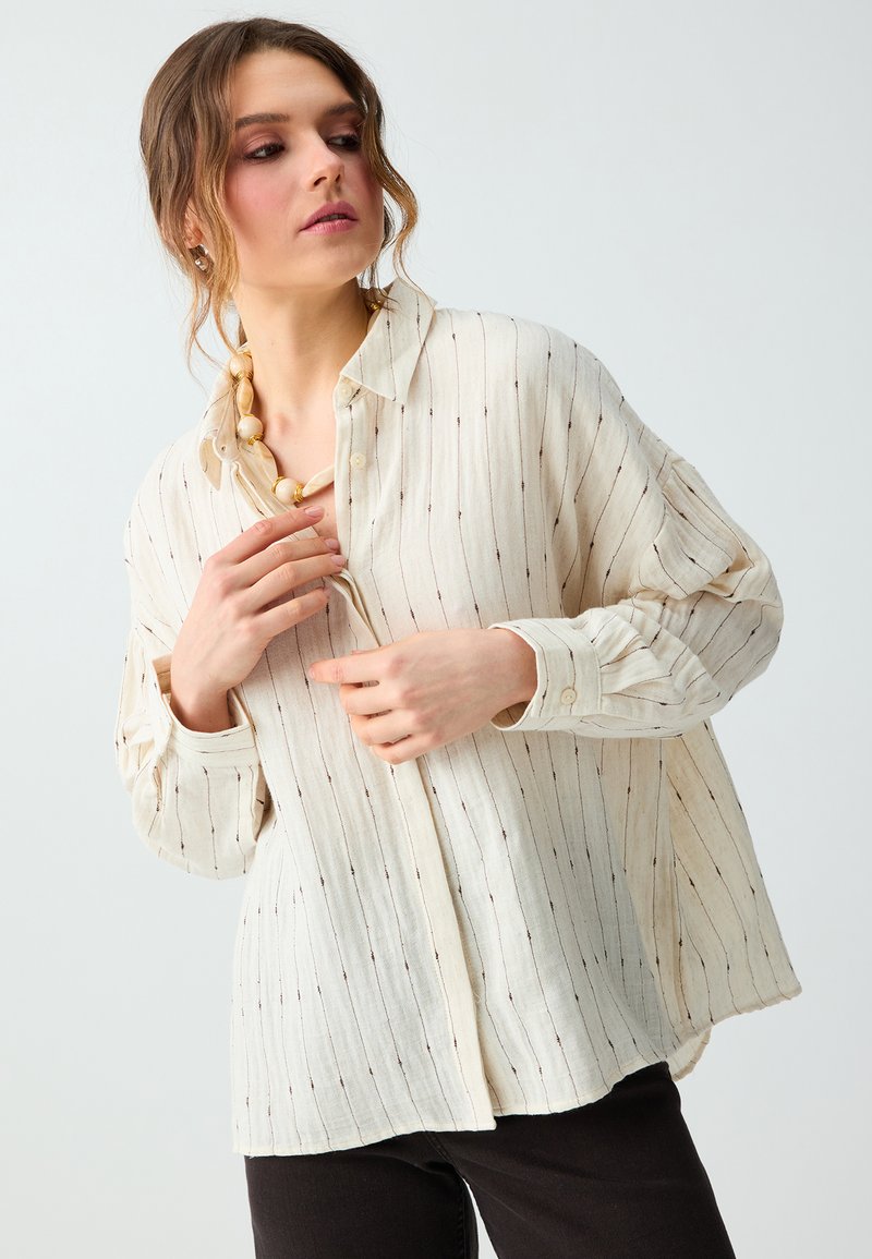 Femme portant une chemise ample crème à manches longues, boutonnée, avec de fines rayures verticales et un collier à grosses perles en bois.
