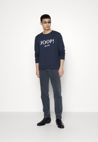 Felpa blu navy con la scritta "JOOP! JEANS" stampata in bianco, abbinata a jeans grigi e stivaletti neri, su uno sfondo bianco uniforme.