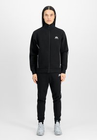 Zwarte sportieve trainingspak gemaakt van zachte stof, met een zipjack in combinatie met witte accenten en bijpassende broek. Draagt lichtgrijze sneakers.