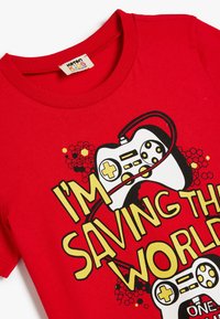 T-shirt in cotone rosso con un design grafico di controller di gioco bianchi e la scritta "STO SALVANDO IL MONDO" in giallo, nero e rosso.