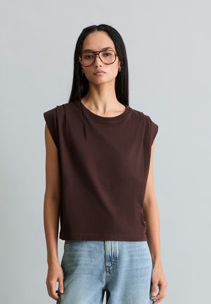 Mujer con cabello largo y oscuro y gafas, vestida con una camiseta sin mangas marrón oscuro y vaqueros azul claro sobre un fondo liso y claro.