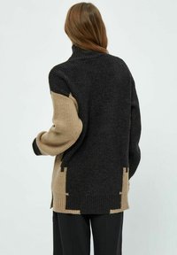 Henkilö, jolla on päällään musta-beige värilohkottu neule, jossa on korkea kaulus, kuvattuna selkäpuolelta yksinkertaista taustaa vasten.