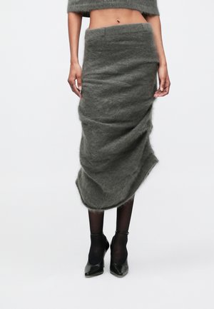 Donna che indossa una gonna midi grigia a vita alta e pelosa, collant neri trasparenti, tacchi neri e un maglione grigio corto su uno sfondo bianco.