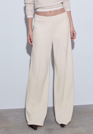 Weite Hose aus weichem, beigem Stoff mit vertikalen Nahtdetails und hoher Taille. Sie bietet eine glatte Textur und eine verlängerte Silhouette.