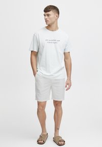 Lyseblå t-shirt med korte ærmer og sort tekst, "HAR DU VANDET DINE PLANTER I DAG?", parret med hvide shorts og brune sandaler.